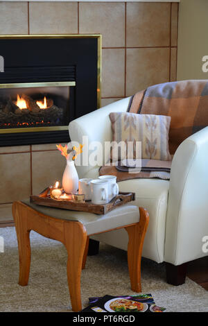 Hygge home Komfort mit weichem Leder bucket Stuhl, Kerzen, Decke und Tee neben Gas Kamin. Vertikales format in natürlichem Licht Stockfoto
