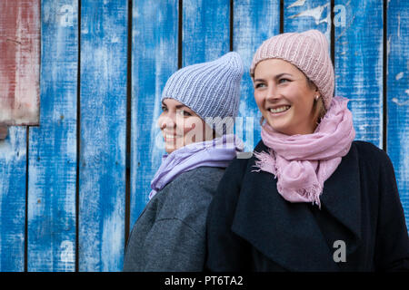 Porträt einer Zwei schöne junge Modelle in Blau und Rosa gestrickte Hüte in der Nähe der blauen Wand stehend auf einem Herbst Tag. Herbst warm Foto. Frau lächelnd und Stockfoto