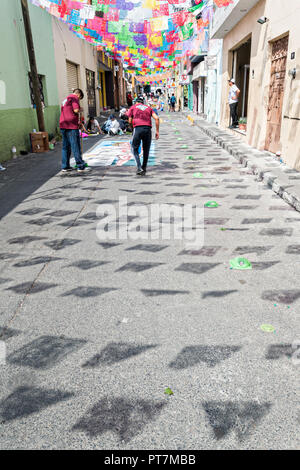 Papel picado Banner werfen einen Schatten auf der Straße als Bewohner schaffen Riesige florale Teppiche aus farbigen Sägemehl hergestellt und mit Blumen während der 8. Nacht Feier gestaltet markiert das Ende des Festes des Hl. Michael in der mexikanischen Stadt Uriangato, Guanajuato. Jedes Jahr schmückt die Stadt 5 km Fahrt mit religiösen Symbolen in der Vorbereitung für die Statue der Schutzheiligen durch die Stadt zu vorgeführt werden. Uriangato wurde eine internationale Sensation nach wowing Brüssel mit Ihren geblümten Teppich auf dem Brüsseler Grand-Place während der Belgien geblümten Teppich Festival angezeigt. Stockfoto