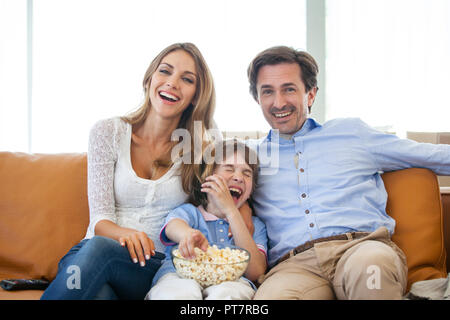 Schöne junge Eltern und deren Sohn sind Fernsehen, essen Popcorn und lächelnd, während auf der Couch zu Hause sitzen Stockfoto