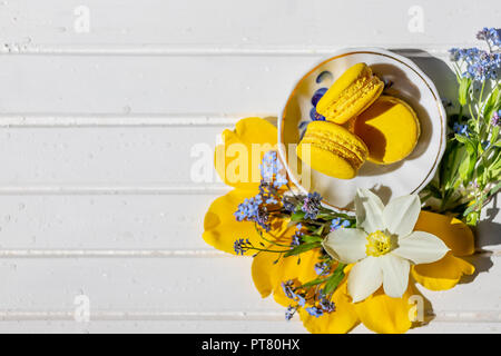 Schönes ruhiges Leben mit Macaron Kekse und Blumen. Zitronen Dessert isoliert. Platz kopieren. französische Schokolade Wüste auf weiße Holztisch. leckeren Nachtisch flach und Narzissen und Vergißmeinnicht Blumen. Stockfoto