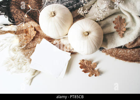 Herbst gemütliche Komposition. Eine leere Karte mockup Szene. Weißen Kürbisse, eiche Blätter, Weihnachtsbeleuchtung und Wolle plaid auf weißem Hintergrund. Erntedank, Herbst, Halloween Konzept. Flach, Ansicht von oben. Stockfoto