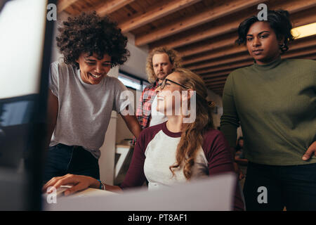 Multi-ethnische Startup-Teams arbeiten im Büro zusammen. Frau, die an ihrem Schreibtisch sitzt und lacht, während das Team dasteht. Business-Team mit ungezwungener Stockfoto