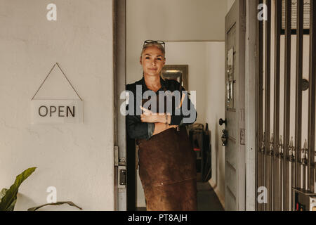 Portrait von älteren weiblichen Goldschmied stehend an Workshop Tür. Frau Juwelier tragen Schürze und Werkzeuge in der Werkstatt. Stockfoto