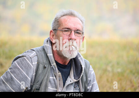 Porträt von einem alten Mann mit grauem Bart und Brille sitzen auf Gras und Rauchen Zigarette Stockfoto