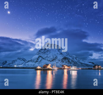 Fischerboote in der Nähe von Pier am Meer gegen Snowy Mountains und starry lila Himmel mit Mond in der Nacht auf den Lofoten Inseln, Norwegen. Winterlandschaft mit Stockfoto