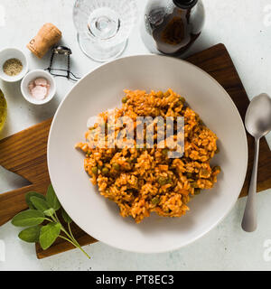 Italienische Risotto mit Tomatensauce und grünen Erbsen, serviert auf einem Teller auf einem hölzernen Schneidebrett und einer Flasche Sekt. Klassische mediterrane Stockfoto