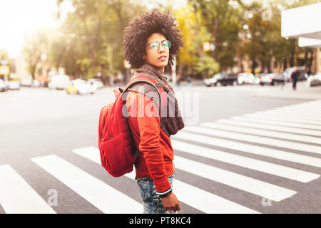 Eine junge afrikanische amerikanische Mann mit rotem Rucksack überquert die Straße an einem fußgängerüberweg in der Stadt. Ein Student wartet, die Straße zu überqueren. Anhand von quantitativen Simulatio Stockfoto