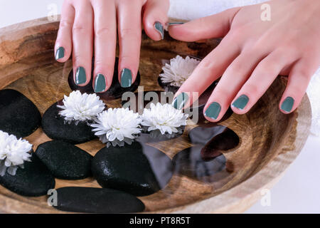 Frau Hände mit grünen Oliven Farbe Maniküre auf Fingernägeln über hölzerne Schüssel mit Wasser und weiße Blume und schwarz Deko Steine. Nahaufnahme Stockfoto