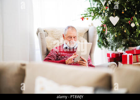 Ein Porträt der ältere Mann mit Tablette auf Sessel zu Hause sitzen zur Weihnachtszeit. Stockfoto