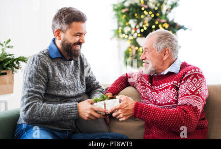 Ein älterer Vater und erwachsener Sohn mit einer gegenwärtigen Sitzen auf einem Sofa in der Weihnachtszeit. Stockfoto