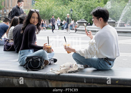 Ein junger Mann in einem Rüschenpullover macht ein Handyfoto einer schönen asiatischen jungen Dame im Washington Square Park in Greenwich Village, New York. Stockfoto