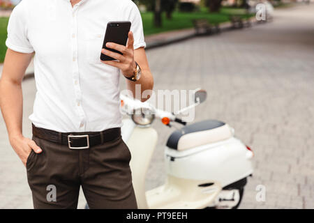 Zugeschnittenes Bild der Geschäftsmann mit Smartphone, während in der Nähe der Roller draußen stehend Stockfoto