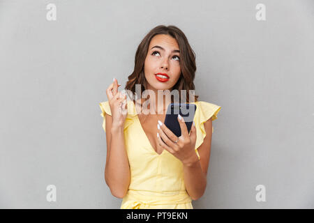 Porträt einer besorgt junges Mädchen im Kleid stehen über grauer Hintergrund, Mobiltelefon, Daumen drücken Stockfoto