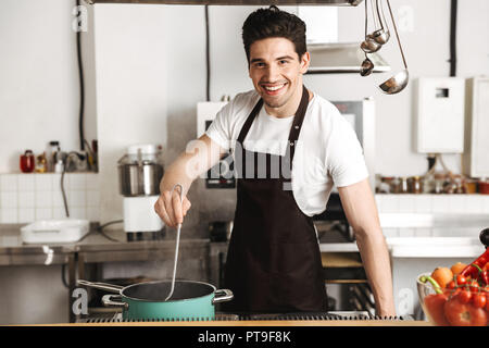 Glückliche junge Mann Chefkoch in Schürze steht in der Küche und rühren, Suppe Stockfoto
