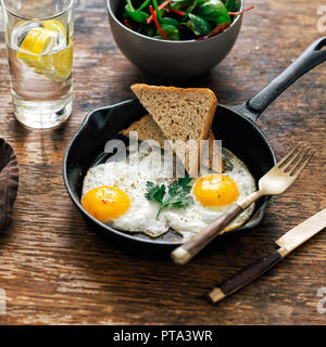 Frühstückstisch. Gebratenes Huhn Eier in der Pfanne mit Brot auf Holztisch mit Salat und Lemon Wasser serviert Stockfoto