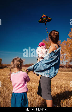 Eine junge Mutter mit ihren zwei Töchtern: ein Kind und einem blonden Mädchen lächeln, Natur und Spiel mit einem Drachen an einem warmen, sonnigen Tag genießen. Das Konzept der Stockfoto