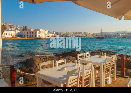 Stühle mit Tischen im typisch griechischen Restaurant in der Altstadt von Mykonos, Kykladen, Griechenland. Panoramablick auf die Marina, den Sandstrand und die Ch Stockfoto