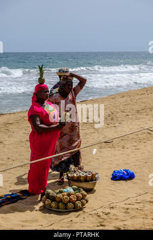 GRAND BASSAM, COTE DIVOIRE - 9. April 2018: Die Frau in der hellen rosa Kleid gleicht einer Ananas auf den Kopf, als sie Früchte für den Verkauf neben Mutter bereitet Stockfoto