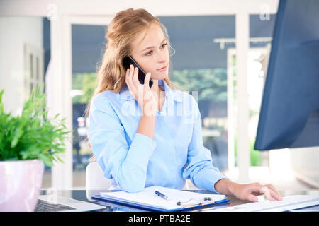 Portrait von schönen jungen Assistenten Geschäftsfrau, Anruf beim Sitzen am Schreibtisch und Arbeiten am Computer. Stockfoto
