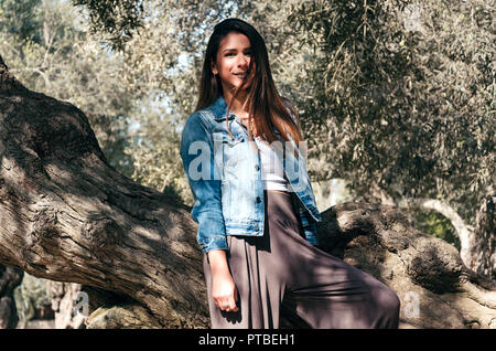 Schöne langhaarige Teenager Frau mit braunen Haaren lehnte sich an einen Baum im Park Stockfoto
