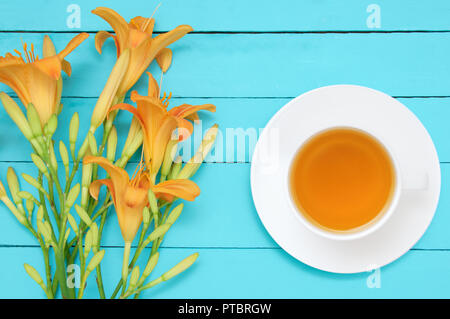 Ein Bouquet von gelben Blumen hemerocallis und eine weiße Schale auf eine Untertasse mit grünem Tee auf einem türkis Holz- Hintergrund. Ansicht von oben Hemerocallis Stockfoto