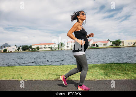 Seitenansicht einer schwangeren Frau auf einem morgendlichen Spaziergang. Frau zu Fuß in der Nähe von einem See Musik zu hören. Stockfoto