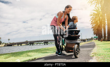 Frau, die ihr Baby in ein Baby Kinderwagen während Ihrer morgendlichen Spaziergang. Junge Mutter mit ihrem Baby in einem Kinderwagen im Park an einem See. Stockfoto