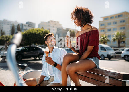Romantisches Paar sitzen auf einer Bank in der Straße essen Kegel Eis am jeweils anderen suchen. Junger Mann und Frau Spaß zu haben, um die Stadt Essen Stockfoto