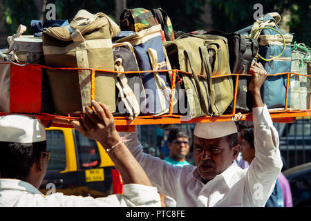 Mit Hilfe eines anderen einen indischen Mann lasten auf ein Korb voller Dabbawala warmes Mittagessen an seinen Kopf um Mumbai, Indien zu liefern. Stockfoto