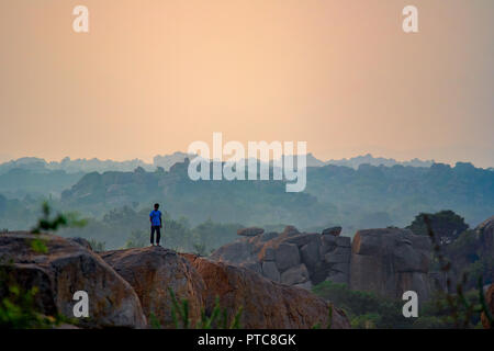 Eine einzige einsame männliche Kletterer steht an der Spitze der beliebten Bouldern Fels mit einer Goldenen Stunde zurück Tropfen Hügel und Felsbrocken. Hampi, Indien. Stockfoto