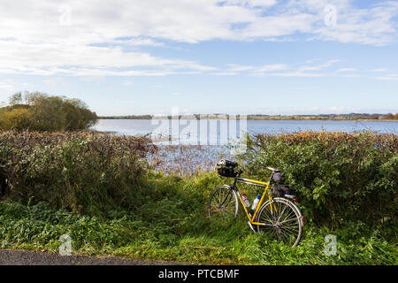 Eine gelbe und schwarze Tourenrad mit vorderen und hinteren Gepäcktasche gegen eine Hecke mit einem weiten Blick über das Wasser. Stockfoto