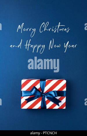 Weihnachtskarte mit Geschenk Box in Rot gestreifte Papier gewickelt und mit blauer Schleife auf dunkelblauem Hintergrund gebunden. Urlaub Konzept, Ansicht von oben. Stockfoto