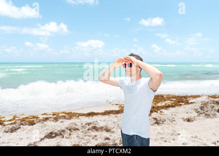 Junger Mann in Sonnenbrille auf Sand, Sandstrand in Miami, Florida, Ozean, Meer, Wasser, Wellen, die an einem sonnigen Tag mit blauem Himmel, Algen, zwei Hände halten Stockfoto