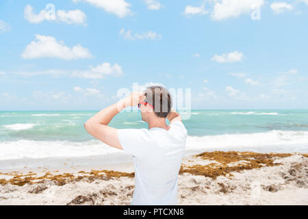 Zurück junger Mann in Sonnenbrille auf Sand, Sandstrand in Miami, Florida, Ozean, Meer, Wasser, Wellen, die an einem sonnigen Tag mit blauem Himmel, Algen, zwei ha Stockfoto