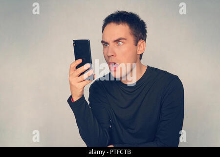 Studio shot junger Mann mit Handy während vor blauem Hintergrund schockiert. Starke Überraschung auf dem Gesicht eines netten Typen Lesen einer Nachricht Stockfoto