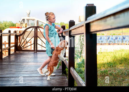 Сhild Mädchen steht Lean in der Nähe der Holzbrücke weg schauen. Hund Welpe Rasse cocker spaniel ruht in der Nähe. Sommer, sonnigen Tag, Abend. Stockfoto