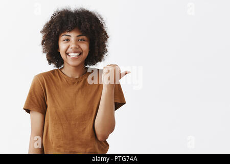 Taille-up Shot von freundlichen, gut aussehende optimistisch dunkelhäutigen Teenager mit Afro Frisur im stilvollen braunes T-Shirt nach rechts mit Daumen und breit grinsend und geben gute Ratschläge Stockfoto