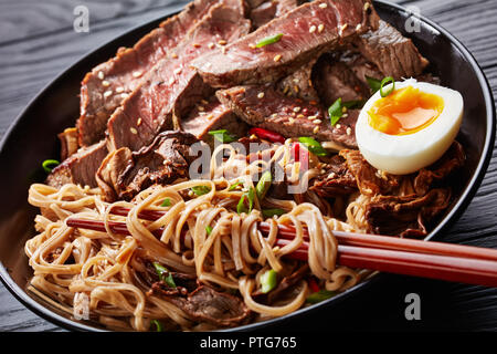 Eine Schüssel mit Soba-nudeln mit in Scheiben geschnittenen Roastbeef Steak und Shiitake Pilze, die Hälfte der hart gekochten Ei und gebratenem Gemüse mit Stäbchen, asiatische Küche, Stockfoto
