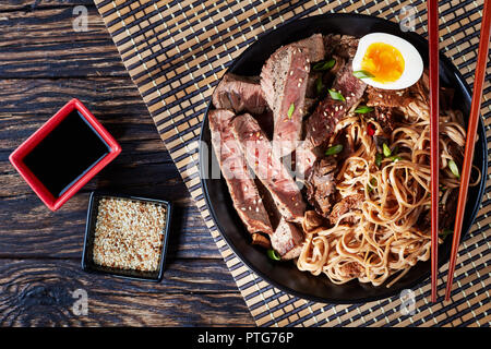 Eine Schüssel mit Soba mit in Scheiben Roastbeef, Shiitake-pilze, die Hälfte der gekochten Ei und Gemüse mit Stäbchen auf einem Bambus Tisch mat. Soja Sauce auf einem Rus Stockfoto