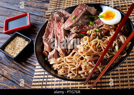 Eine Schüssel mit Soba mit in Scheiben Roastbeef, Shiitake-pilze, die Hälfte der gekochten Ei und Gemüse mit Stäbchen auf einem Bambus Tisch mat. Soja Sauce auf einem Rus Stockfoto