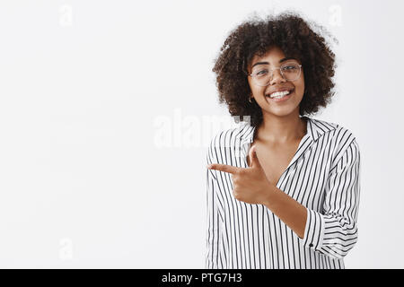 Taille - geschossen von freundlich und höflich, gut aussehende African American Girl in Gläsern mit lockigem Haar in gestreiften Büro Bluse zeigt mit Finger Pistole links und grinsend mit Freude beraten, wo Sie Stockfoto