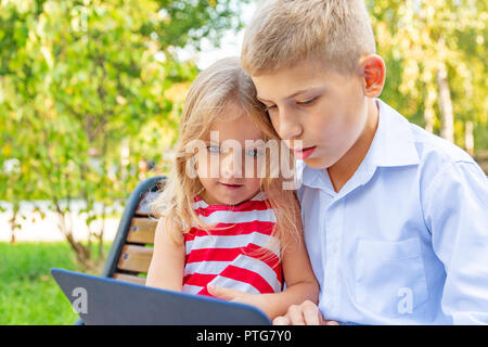 Lächelnd Bruder und Schwester sitzt auf der Bank im Park und spielen auf Laptop