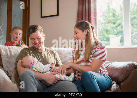 Der Mann und die Frau sitzt auf einem Sofa im Wohnzimmer mit Ihrem neugeborenen Baby. Der Mann hält das Baby und seine Frau versucht, lächelnd und Laug Stockfoto