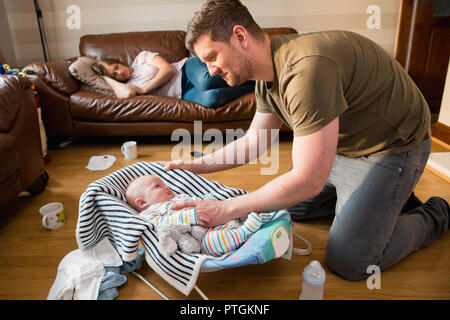 Vater auf sein neugeborenes Baby Boy als er aufwacht aus einem Schlaf und tryig ruhig zu halten, während seine erschöpften Frau hat ein Nickerchen auf dem Sofa. Stockfoto
