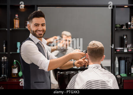 Lächelnd hairstylist arbeiten mit Kunden in Barbershop. Bärtige Friseur an Kamera und Trimmen stilvolle Frisur des jungen Mannes. Reflexion im Spiegel der Client bedeckt mit coiffure Kap. Stockfoto