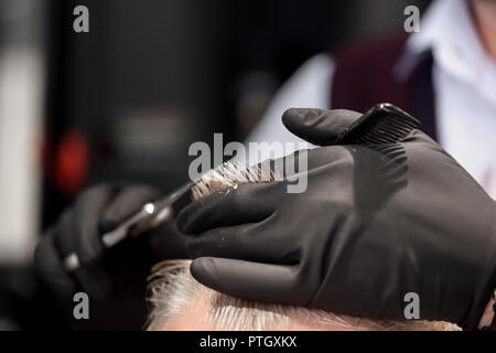 Nahaufnahme des unkenntlich Friseur arbeiten mit Client und Herstellung professioneller Haarschnitt. Hairstylist in speziellen schwarzen Handschuhe aus Kunststoff schwarz Kamm und Schere. Konzept der Barbershop. Stockfoto