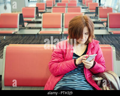 Hübsches Mädchen am Flughafen Lounge sitzen auf Ihrem Smartphone warten auf Flug reise mit kleinen Rucksack. Vorderansicht shot mit viel Platz em kopieren Stockfoto