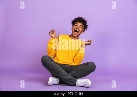 Portrait von Angeregten African American Boy schreien und Suchen zur Seite beim Sitzen auf dem Boden mit gekreuzten Beinen über violett Hintergrund isoliert Stockfoto