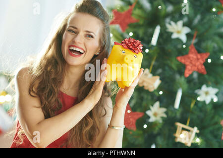 Gerne trendy Frau im roten Kleid schütteln gelbe Sparschwein mit roter Schleife in der Nähe von Weihnachten Baum Stockfoto